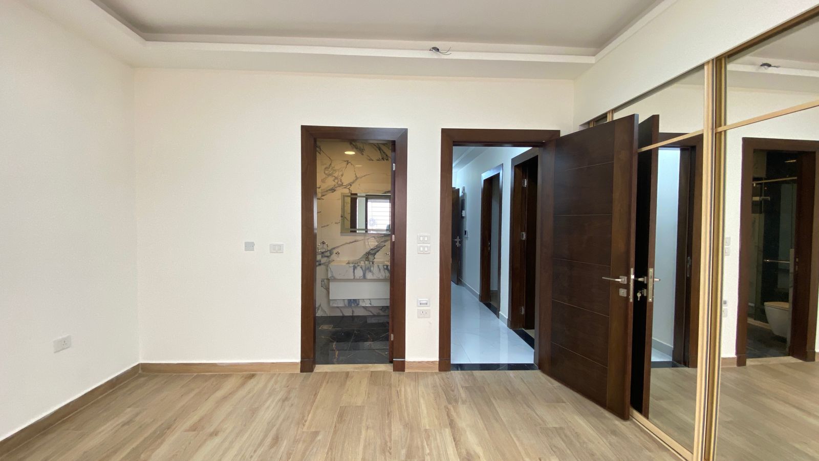 36532989-fcf0-4ef4-a490-ca177f35138a Empty room with light wood flooring and three doorways: one to a marble-floored bathroom, one to a hallway, and a large mirrored wardrobe along the right wall.