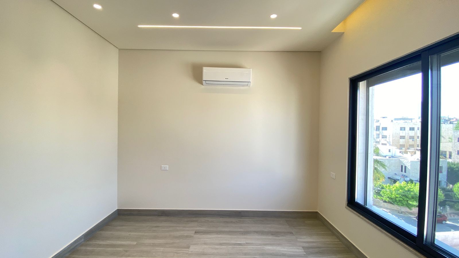 3747194c-df4e-455d-a93d-6c7d60e22560 Empty modern room with light wood floor, white walls, ceiling recessed lights, a wall-mounted AC unit, and a large window showing a city street outside.