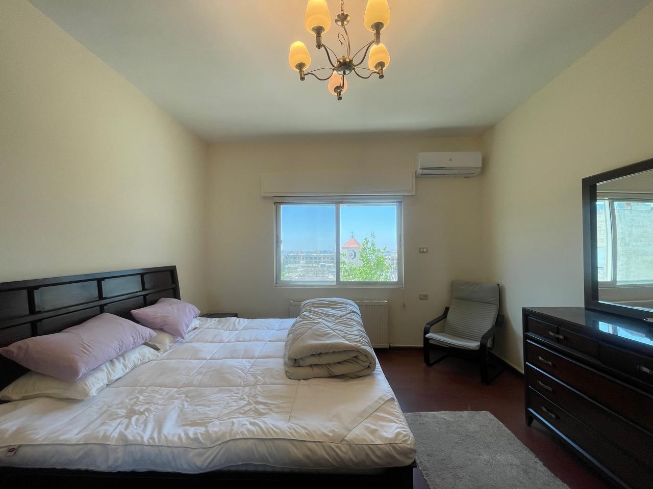 37984362-ed70-4583-a436-a6709254c08a A bedroom with a dark wood bed, white comforter, and pink pillows facing a window with a city view; a rolled gray blanket sits on the bed.