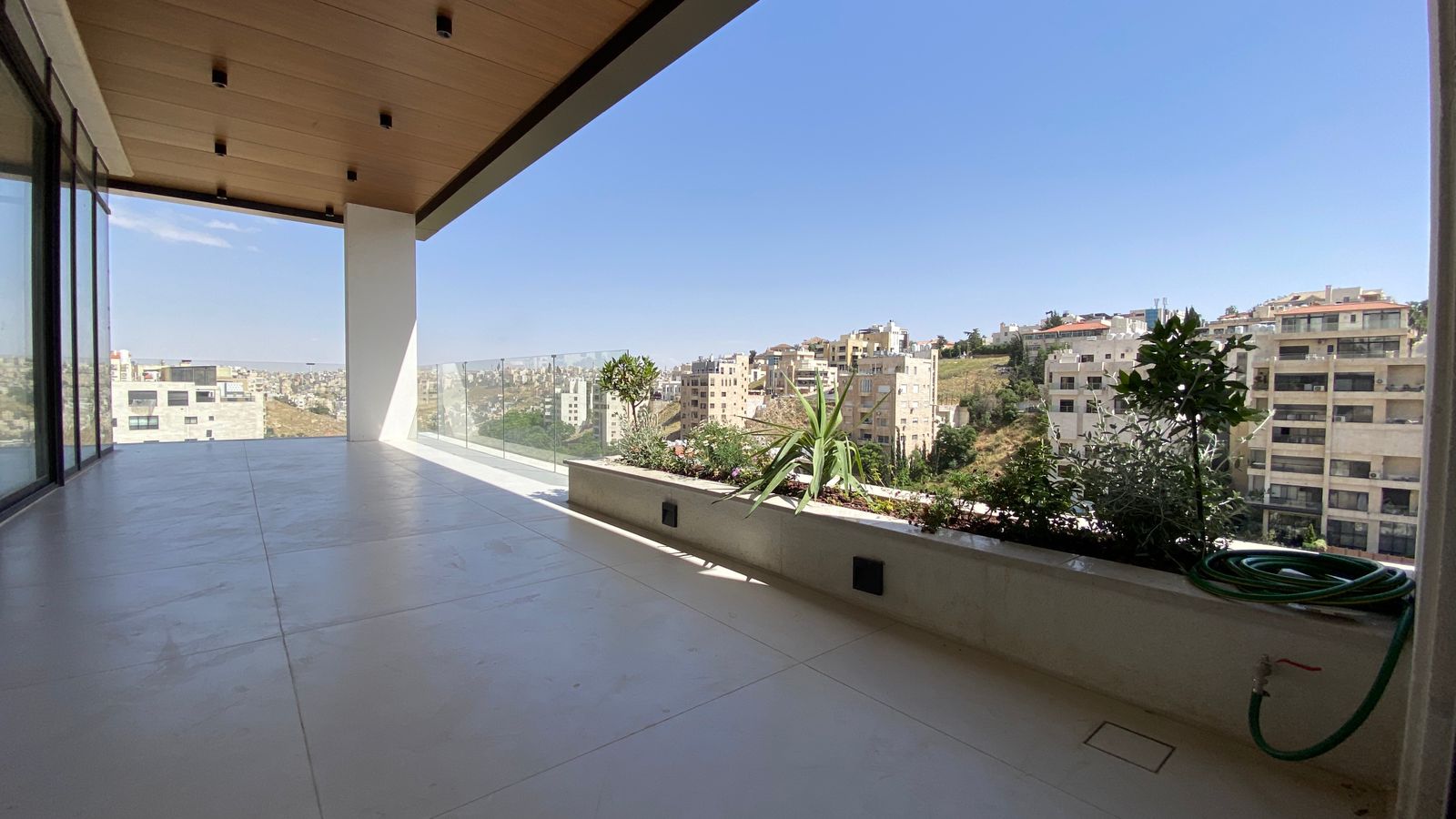 37cfacf2-9bd5-460d-8859-164cd4cfa845 Modern balcony with glass railing overlooking a cityscape and clear blue sky; planters along the edge with plants and a coiled hose nearby.