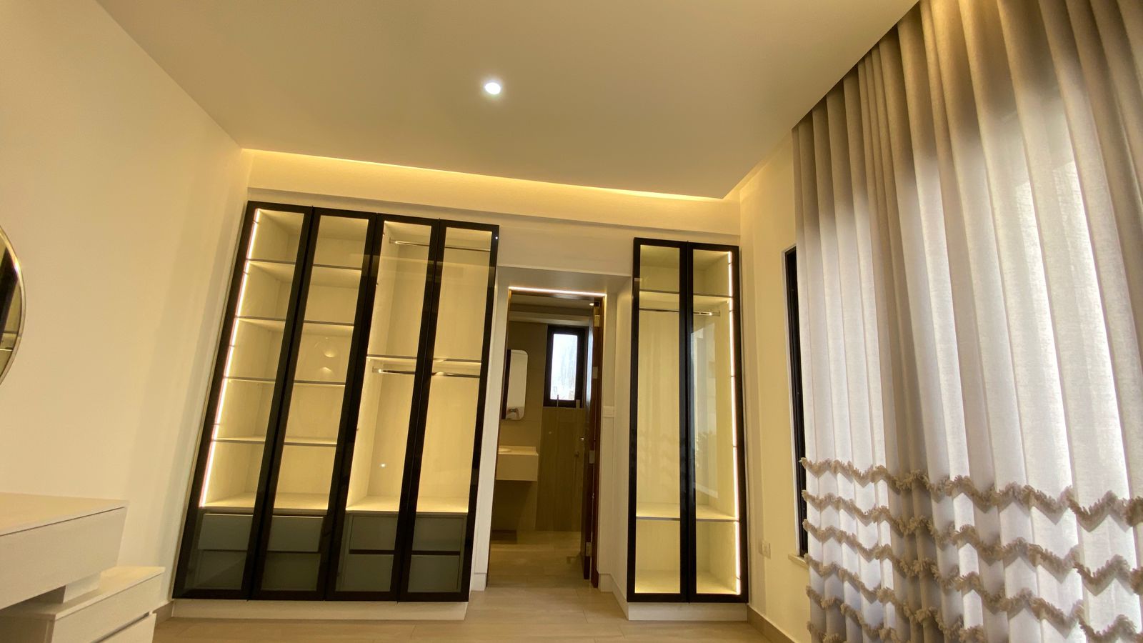 3865aeec-2a27-435b-b860-139a3ff0a2ef Modern walk-in wardrobe with glass-fronted black frames and warm built-in lighting, seen from a bright room with sheer curtains on the right