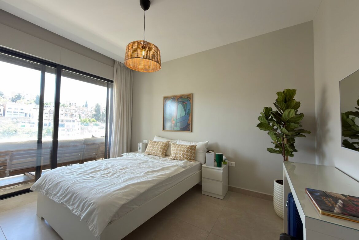 Bright modern bedroom with a white bed, beige pillows, and a nightstand by a large glass balcony door letting in natural light.