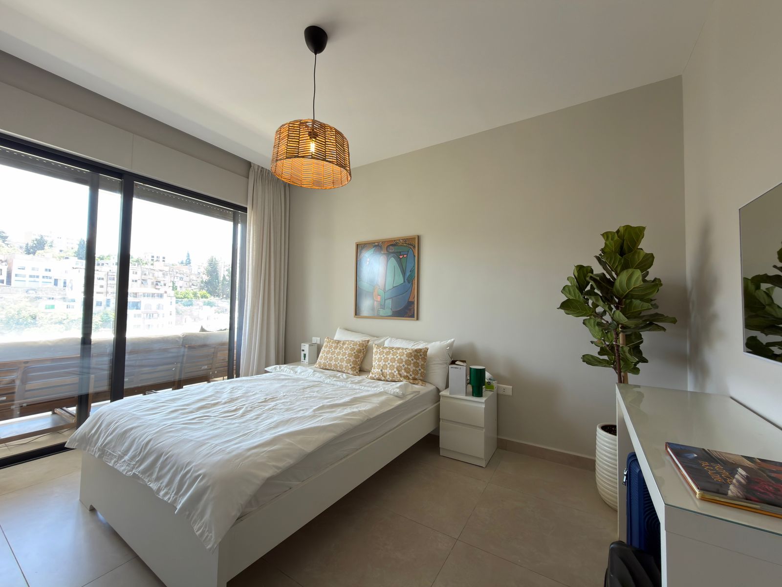 389f0b87-630d-4df4-a53f-ec8a65a5a4c8 Bright modern bedroom with a white bed, beige pillows, and a nightstand by a large glass balcony door letting in natural light.