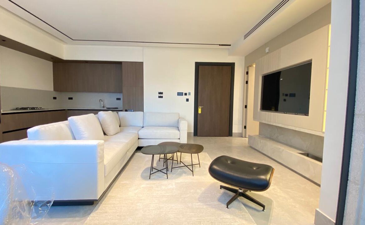 Bright modern living room with a white sectional sofa, small round coffee tables, and a black lounge chair. A kitchen is in the background.