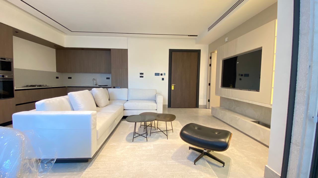 Bright modern living room with a white sectional sofa, small round coffee tables, and a black lounge chair. A kitchen is in the background.