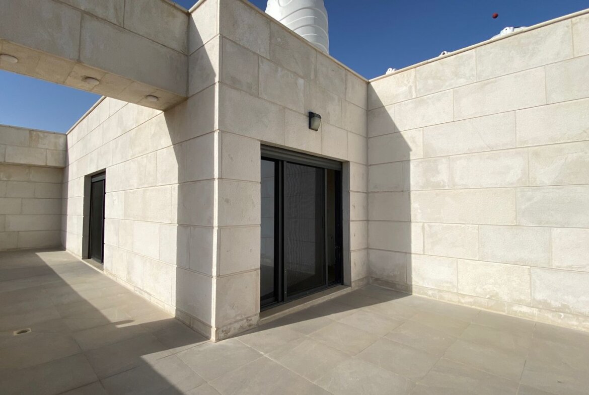 Beige stone courtyard with a sliding glass door and a shaded doorway on the left, under a clear blue sky.