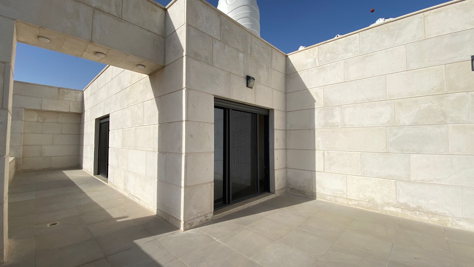 Beige stone courtyard with a sliding glass door and a shaded doorway on the left, under a clear blue sky.