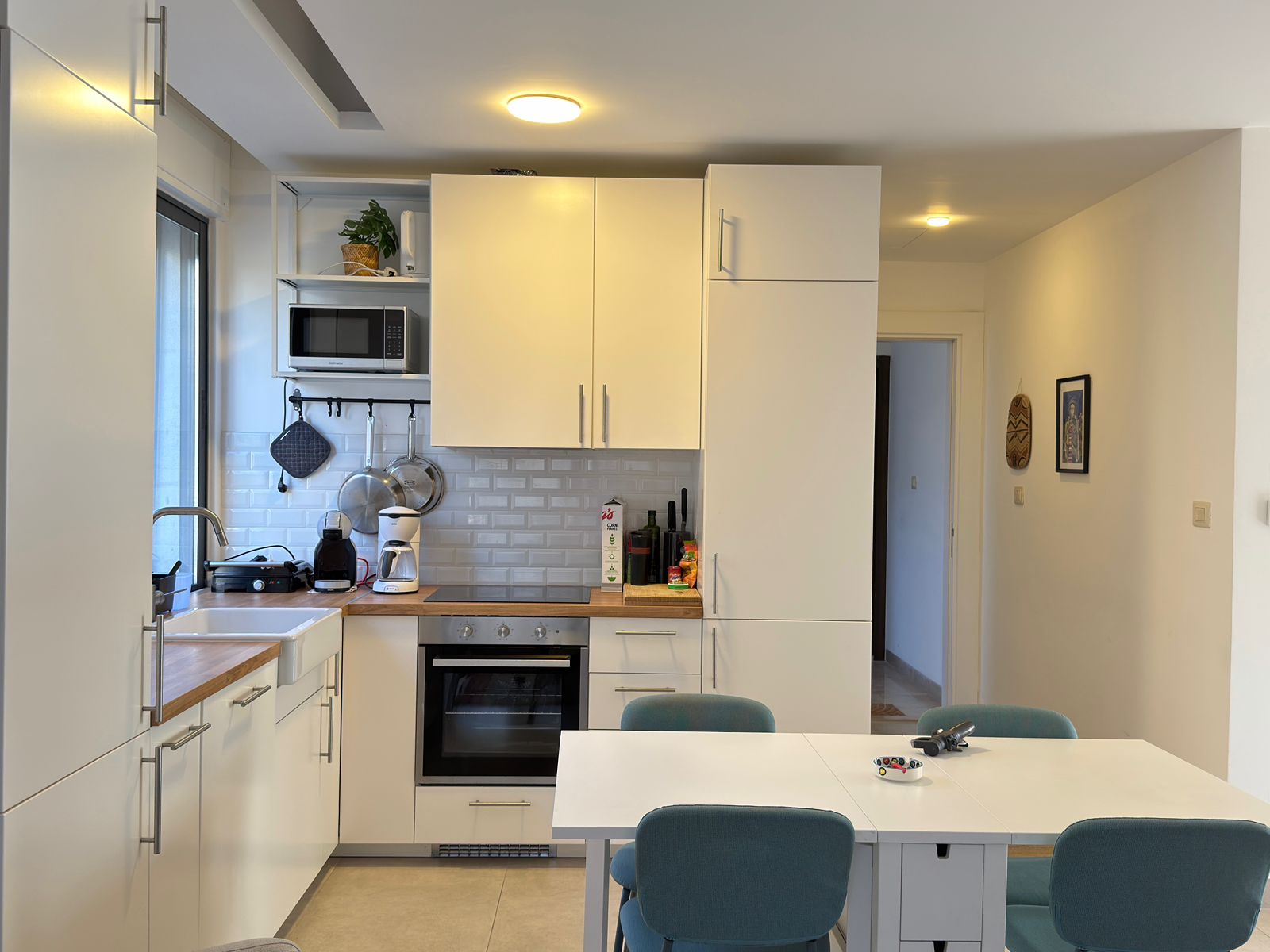 3906178c-7a42-45af-a6a8-adc0aae21ef8 Modern white kitchen with built-in oven, microwave shelf, and a wooden counter against a tiled backsplash; teal chairs around a white dining table in foreground.