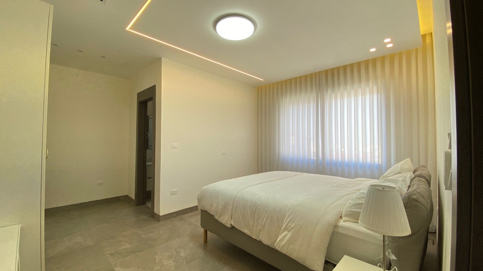 39117b80-28a2-4141-9a3b-65191d46e378 Bright, modern bedroom with a large bed in white linens, sheer curtains, and a bedside lamp.