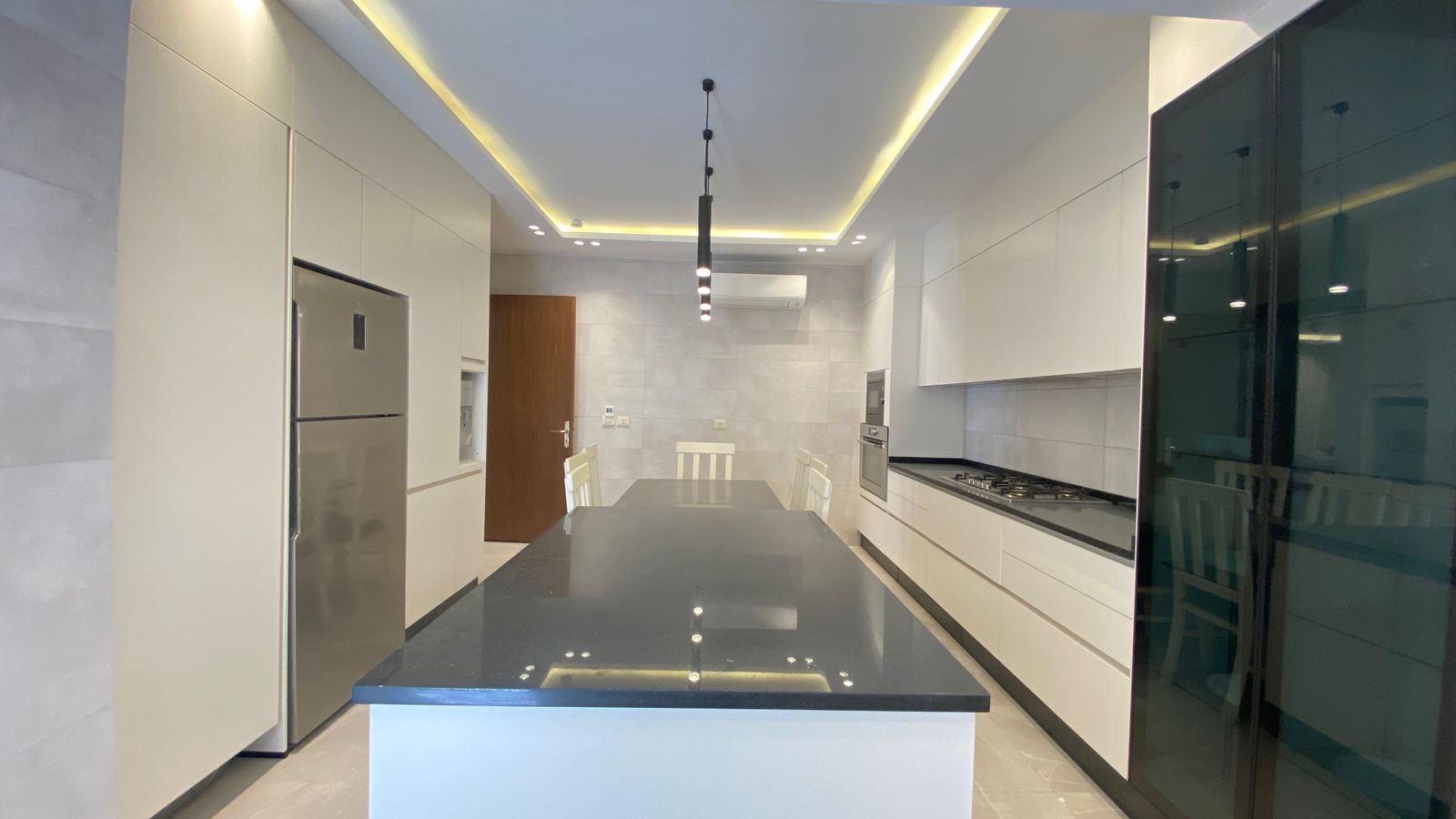 3989c9df-f064-433b-8f74-e7e71a787413 Modern white kitchen with a dark countertop island, stainless steel fridge, and built‑in appliances along the right wall.