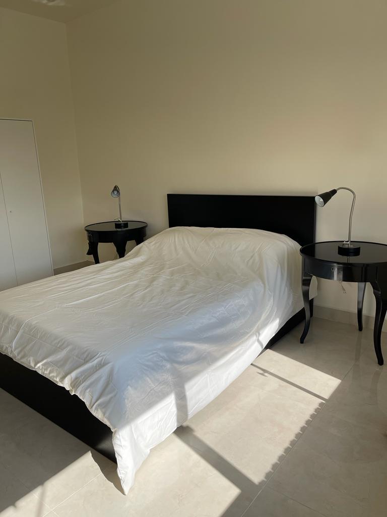39cbccc8-4708-478c-8c2b-bf41805c3f50 Queen-size bed with white duvet and black headboard, flanked by matching black nightstands with lamps in a bright minimalist bedroom.
