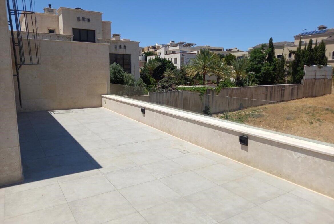 3a0453a6-c6c5-446c-9bbc-969b50facbc9 Sunlit beige tiled terrace with a low glass railing overlooking a dry yard and palm trees in a residential area.|A wide, modern terrace in pale stone tiles under a clear blue sky, with glass railing and neighboring beige buildings in view.