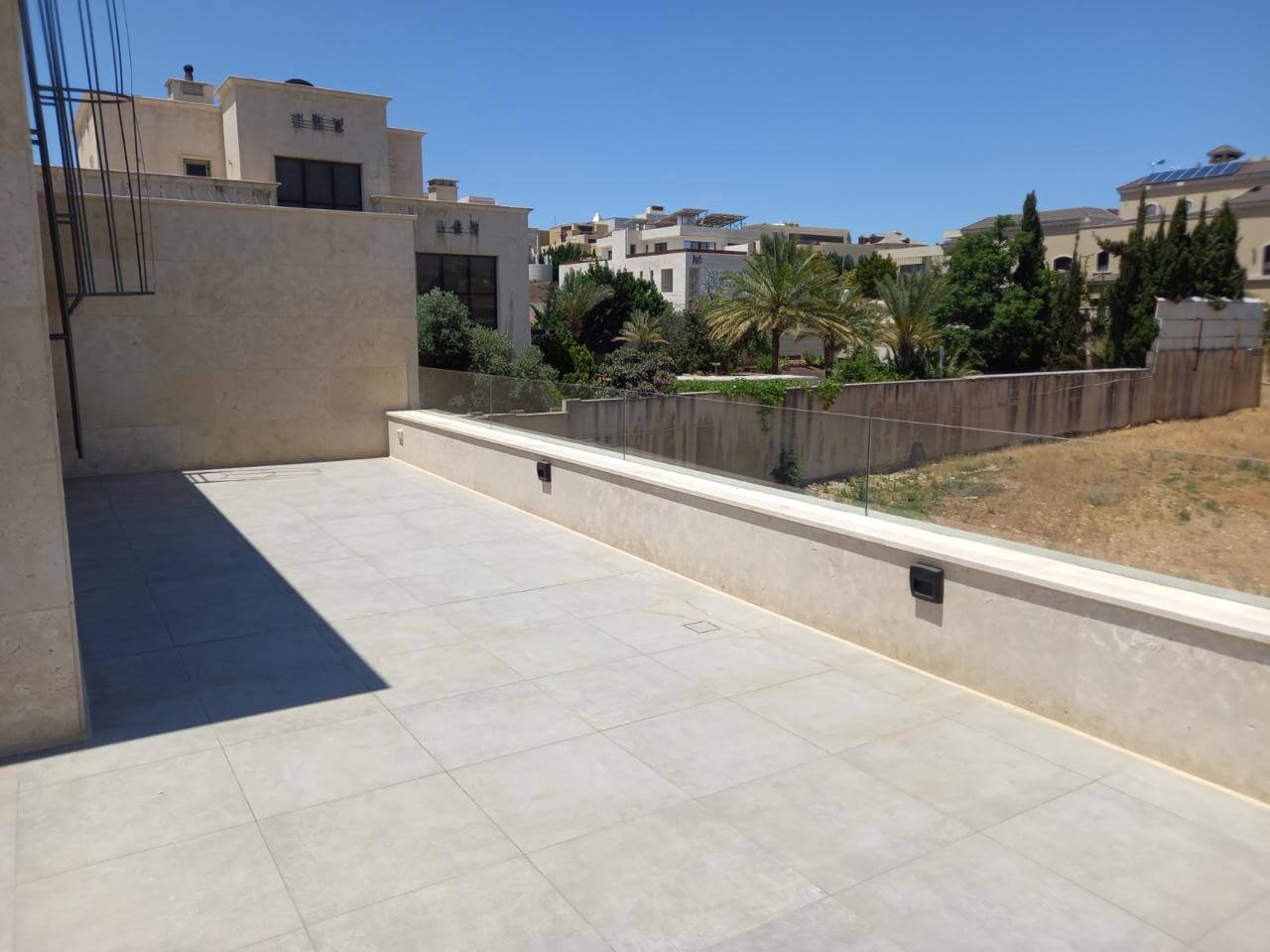 3a0453a6-c6c5-446c-9bbc-969b50facbc9 Sunlit beige tiled terrace with a low glass railing overlooking a dry yard and palm trees in a residential area.|A wide, modern terrace in pale stone tiles under a clear blue sky, with glass railing and neighboring beige buildings in view.
