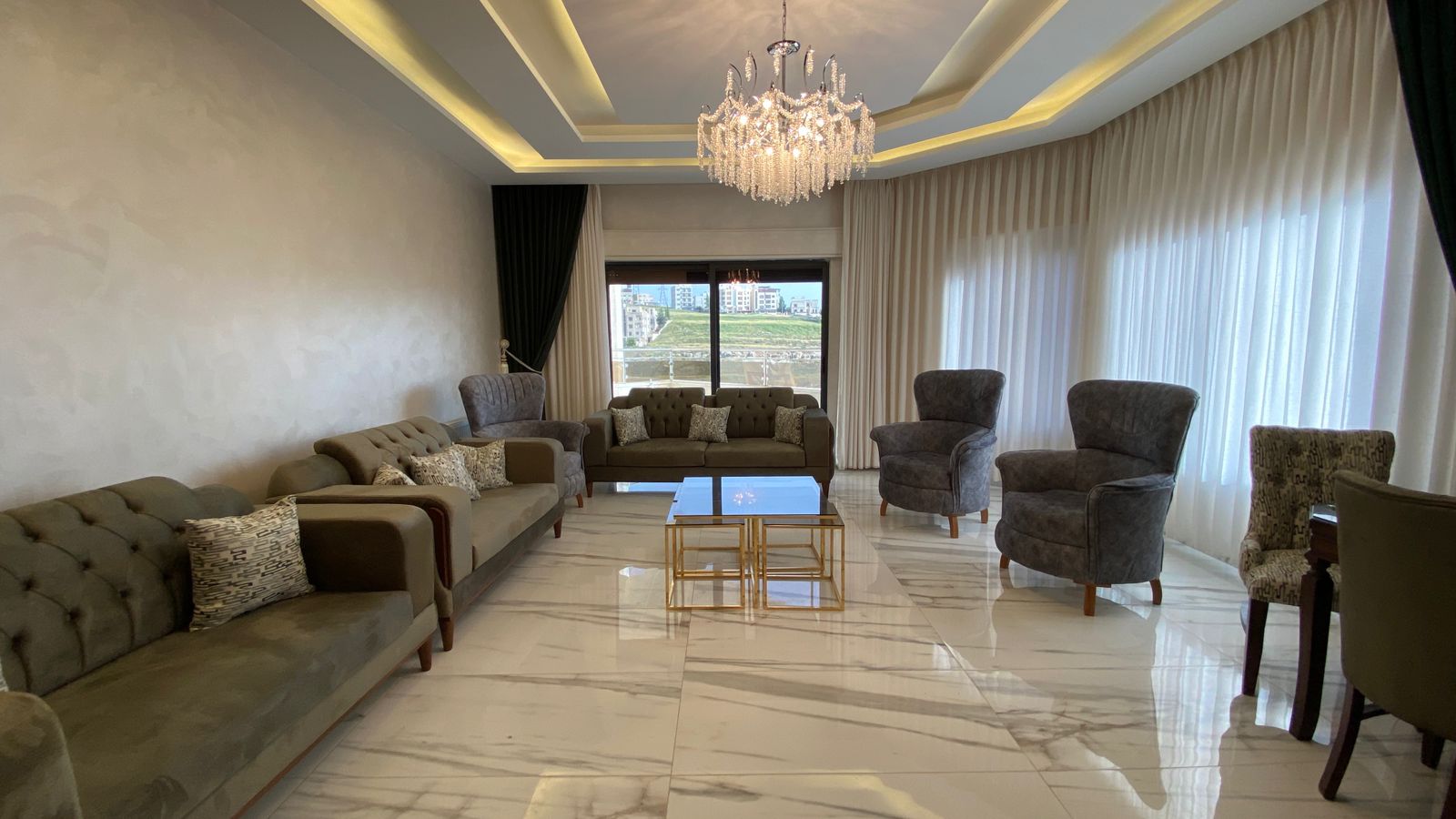 Luxurious living room with marble floor, grey sofas, chandelier, and large windows with sheer curtains.