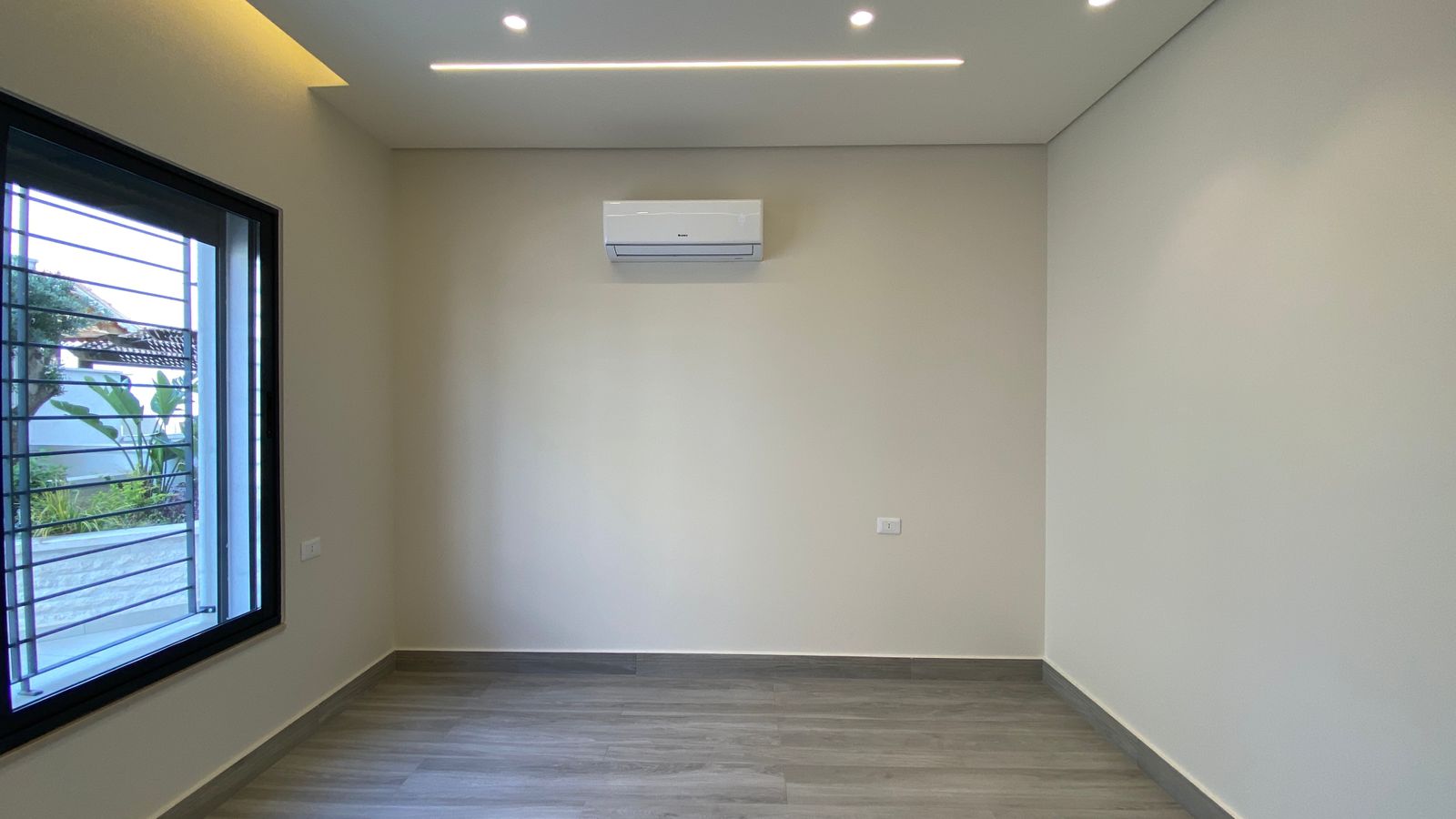 3a7316d4-28e3-4c57-b179-73e588f74750 Empty modern room with a wall-mounted air conditioner, large window on the left, and gray wood-like flooring.