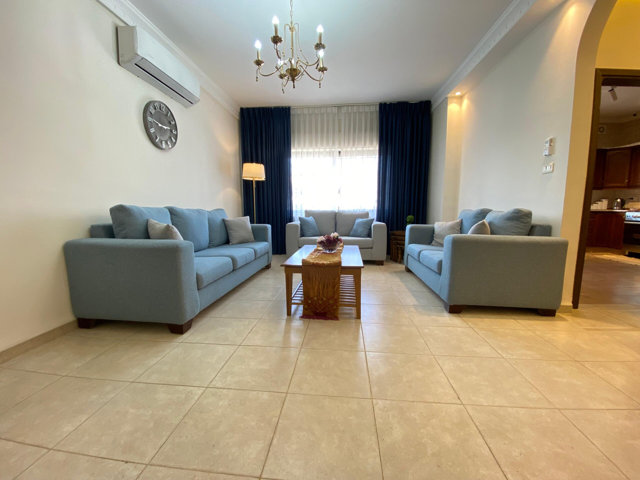 3b07dfdf-6c00-4eb6-8bed-52d4d10359d1 Living room with light blue sofas arranged around a wooden coffee table, cream tile floor, and dark blue curtains framing a bright window. A chandelier hangs from the ceiling, and an air conditioner is mounted on the wall. A clock is visible on the left wall.