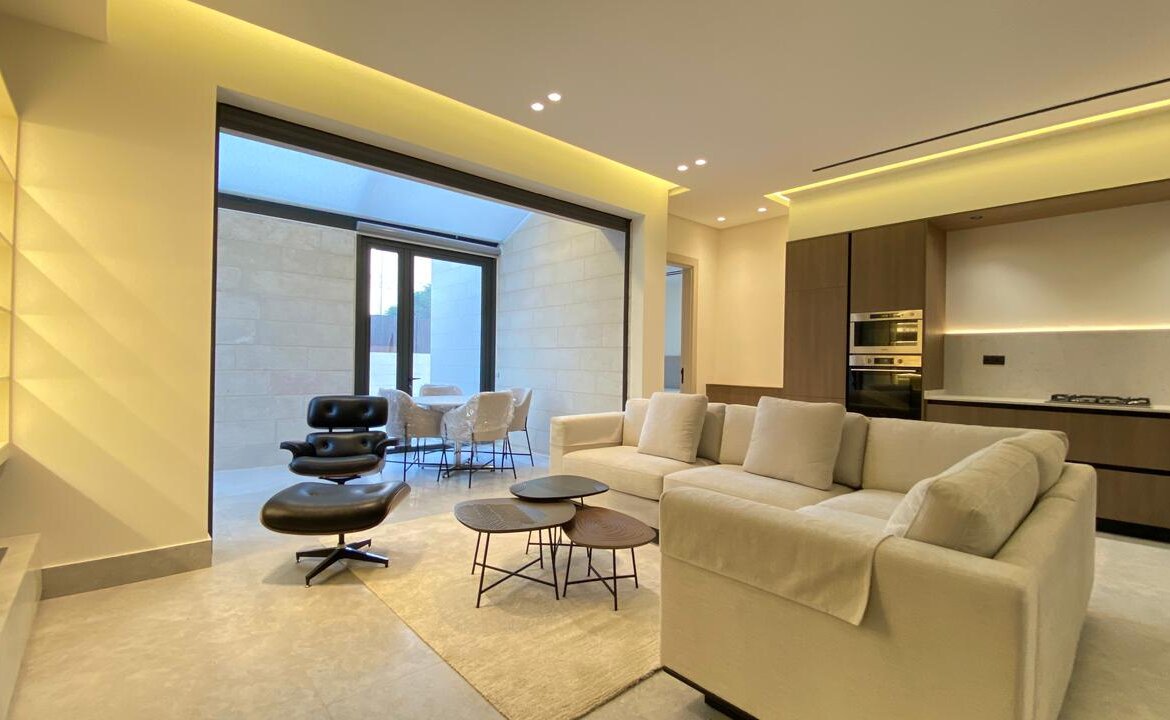 Bright open-plan living room with a cream sectional, black lounge chair with ottoman, and a set of round coffee tables near a glass patio door to an outdoor seating area.