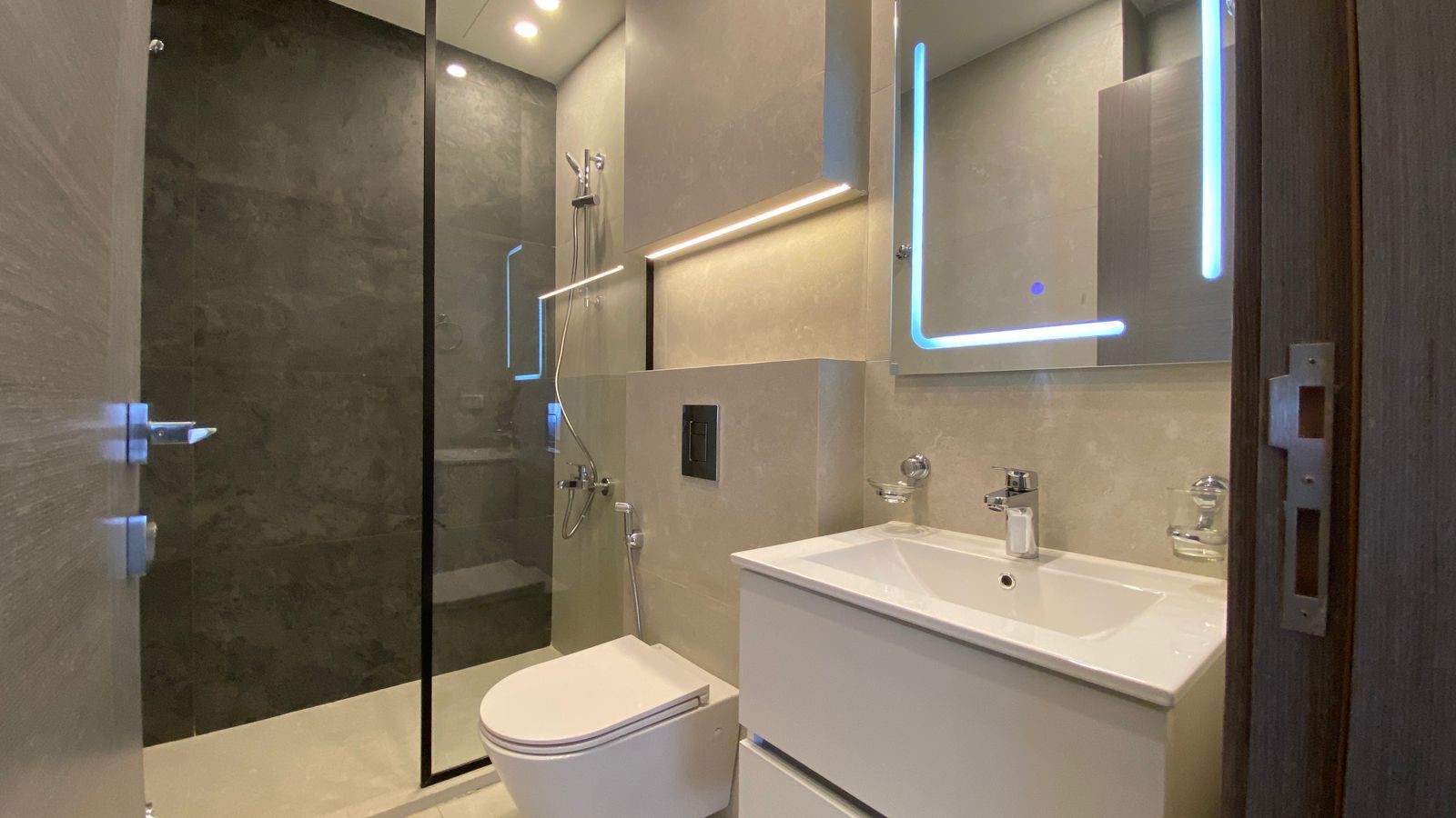 3bcbc0e3-aed6-4930-97a3-4c7b078cf864 Modern bathroom featuring a glass-enclosed shower, wall-mounted toilet, and a white vanity with sink and backlit blue-LED mirror.