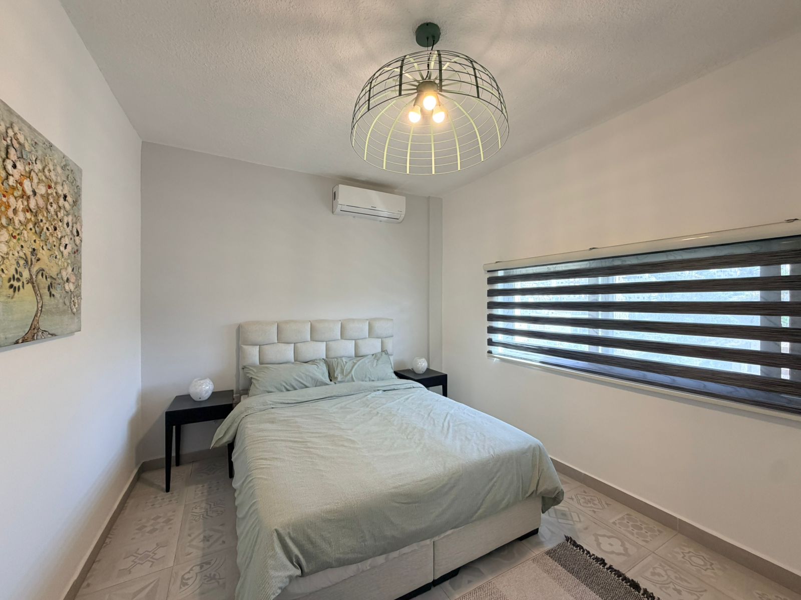 3c4b959e-fa4f-4ff7-a25c-919f1927ff68 Bedroom with a padded gray headboard, mint bedding, two black nightstands, and a geometric ceiling light above the bed.