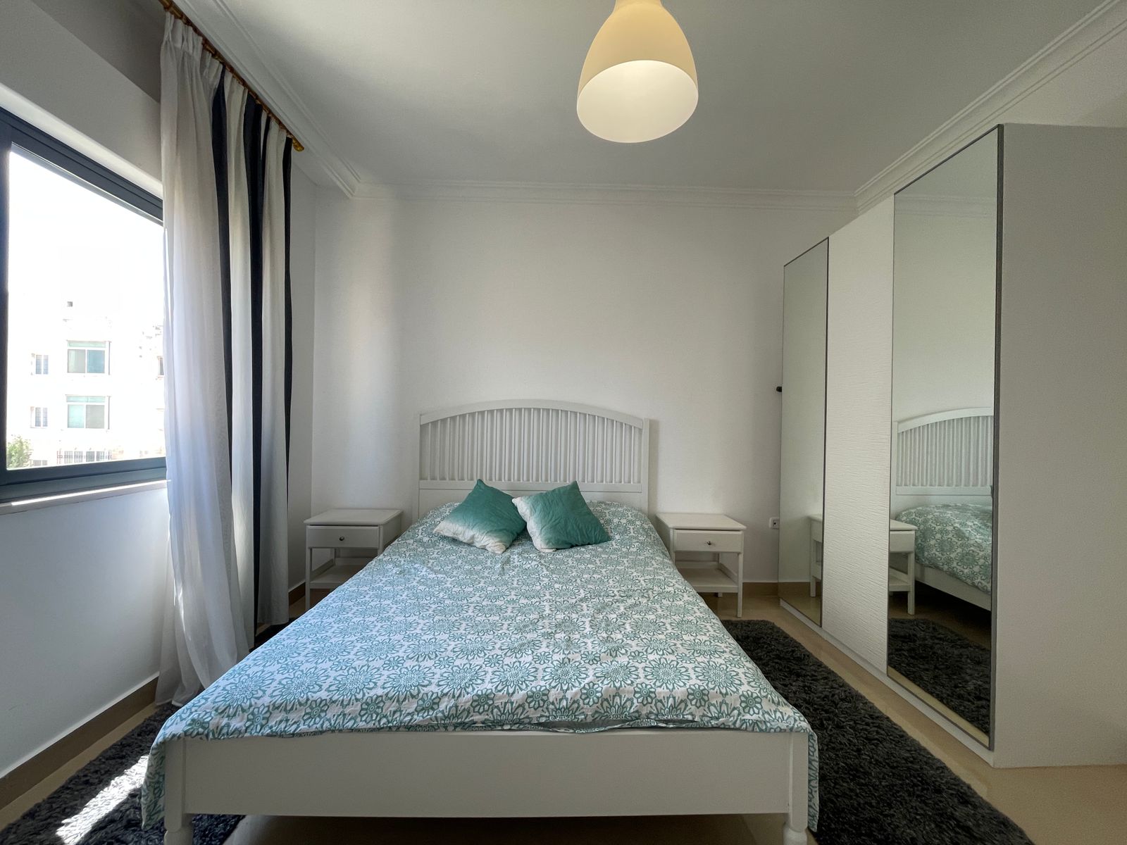 3c51e1d2-0811-4605-a206-598e8a71b4f7 Bright white bedroom with a white wooden bed, teal patterned bedding, and two white nightstands under a large window with sheer black-and-white curtains and a pendant light overhead