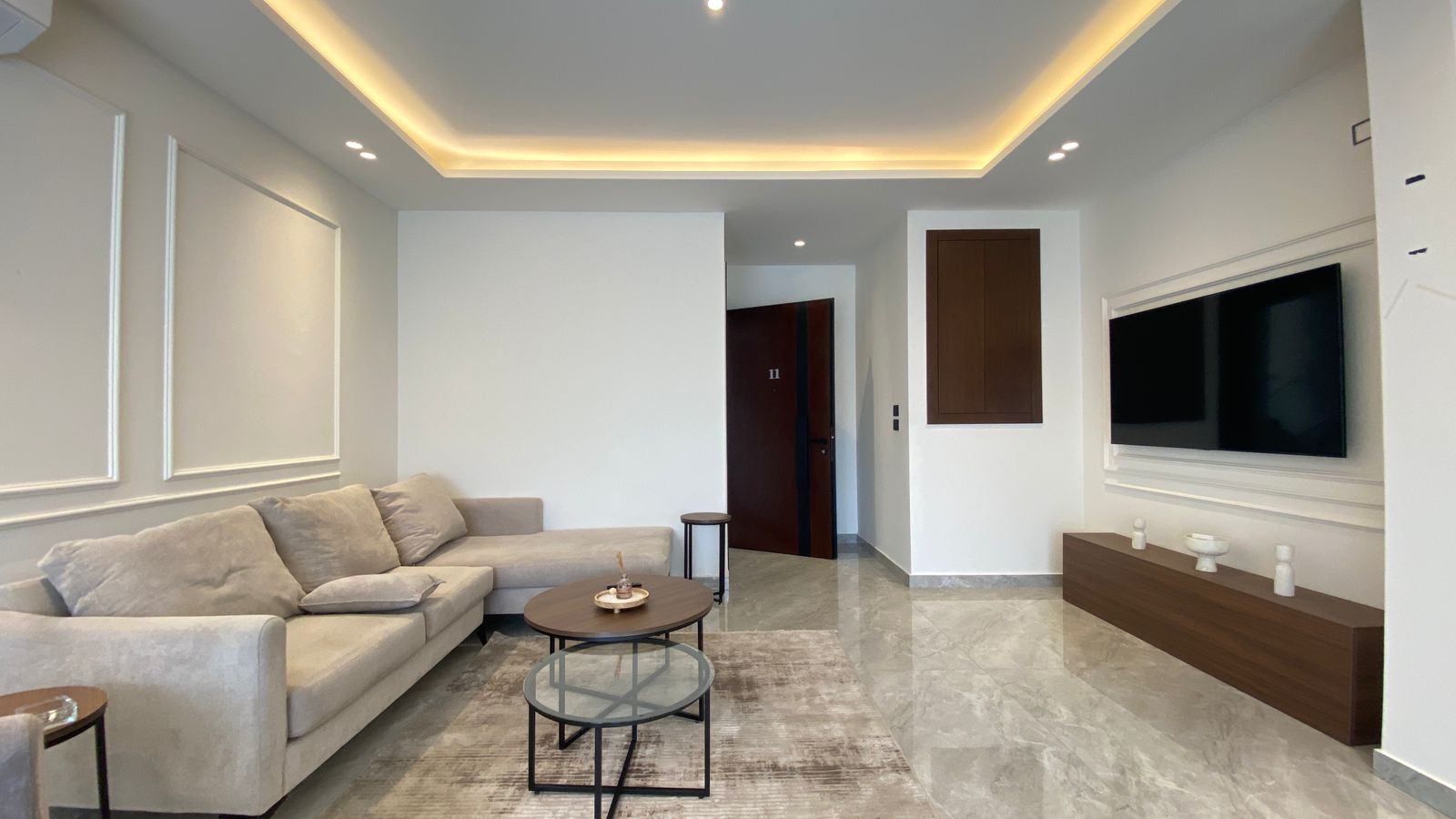 3c619404-1795-460a-8ee8-ef3414dfa2de Modern living room with a light beige sectional sofa, round coffee tables, and a textured rug under a TV wall and low wood console.