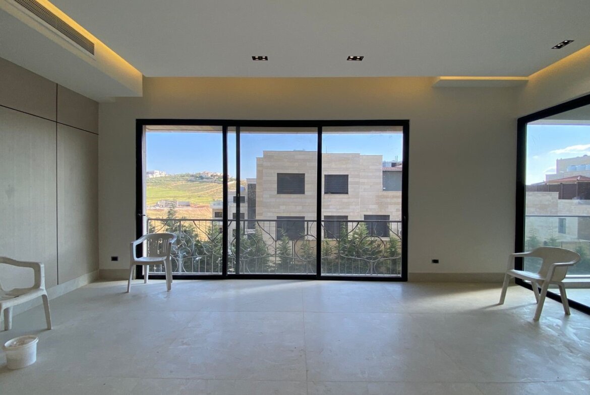 Bright, unfurnished room with large sliding glass doors opening to a balcony; three white plastic chairs and a bucket on the pale floor.