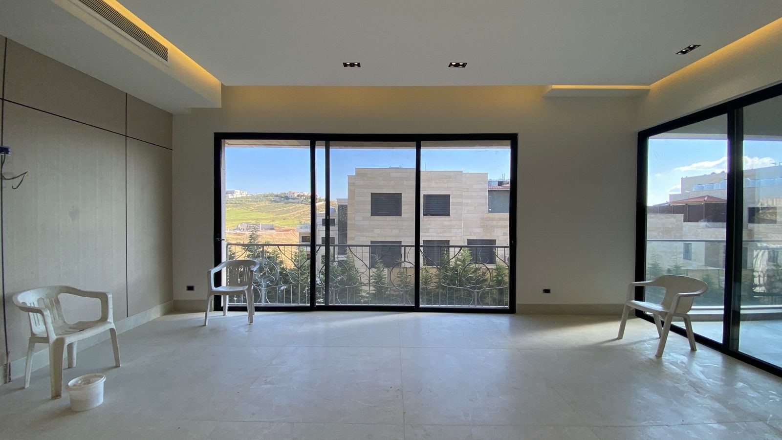3c97f7c9-34c7-4344-b698-47e4b5da2809 Bright, unfurnished room with large sliding glass doors opening to a balcony; three white plastic chairs and a bucket on the pale floor.