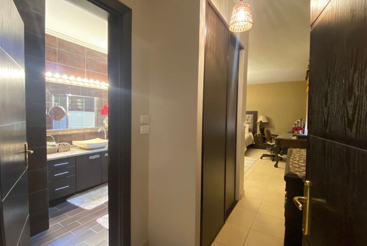 3ca4e324-e53c-4263-a763-edd03d71540a Hallway with an open bathroom on the left showing a lit vanity mirror and sink; hallway leads to a bedroom/office area in the distance.
