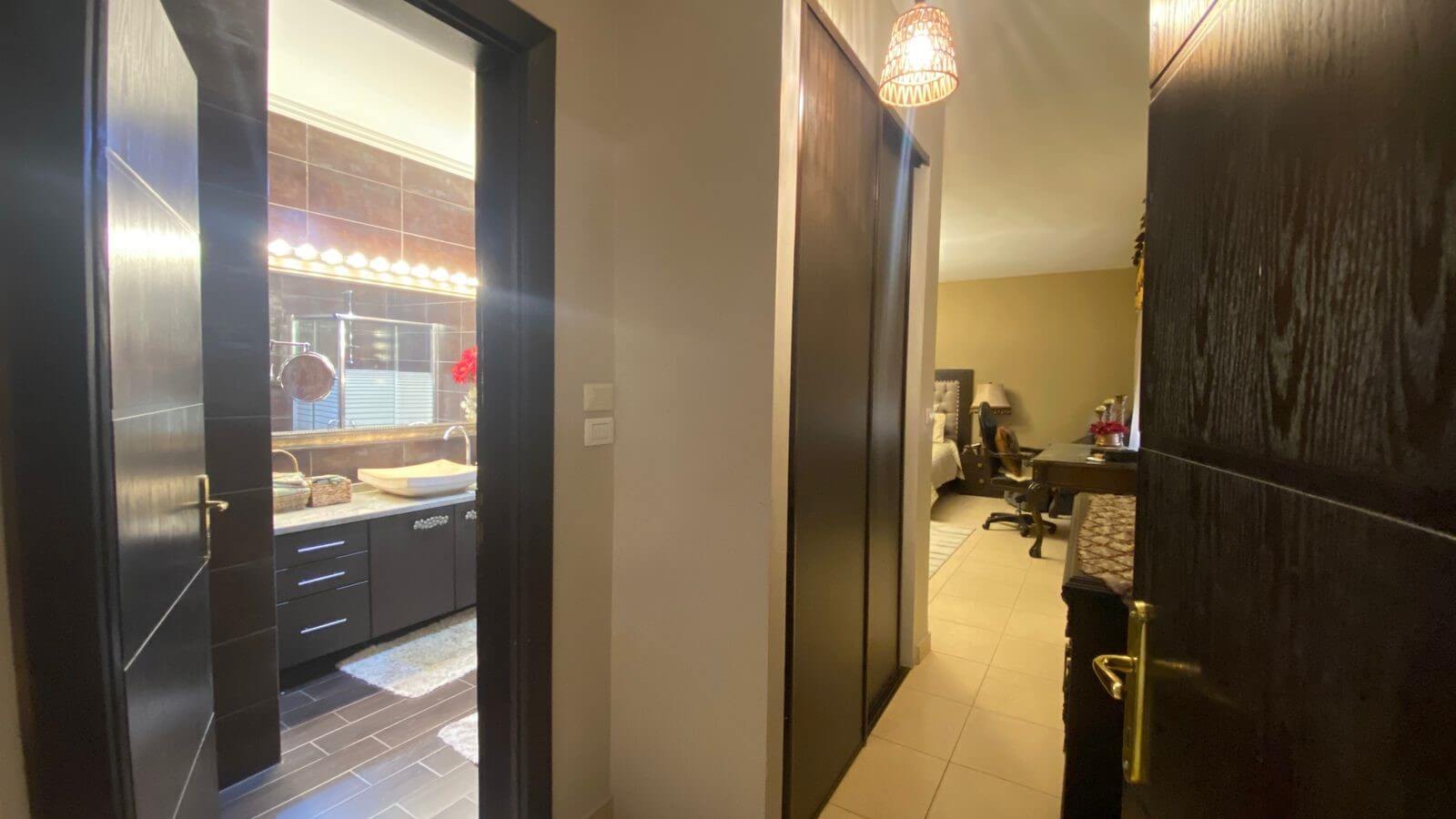 3ca4e324-e53c-4263-a763-edd03d71540a Hallway with an open bathroom on the left showing a lit vanity mirror and sink; hallway leads to a bedroom/office area in the distance.