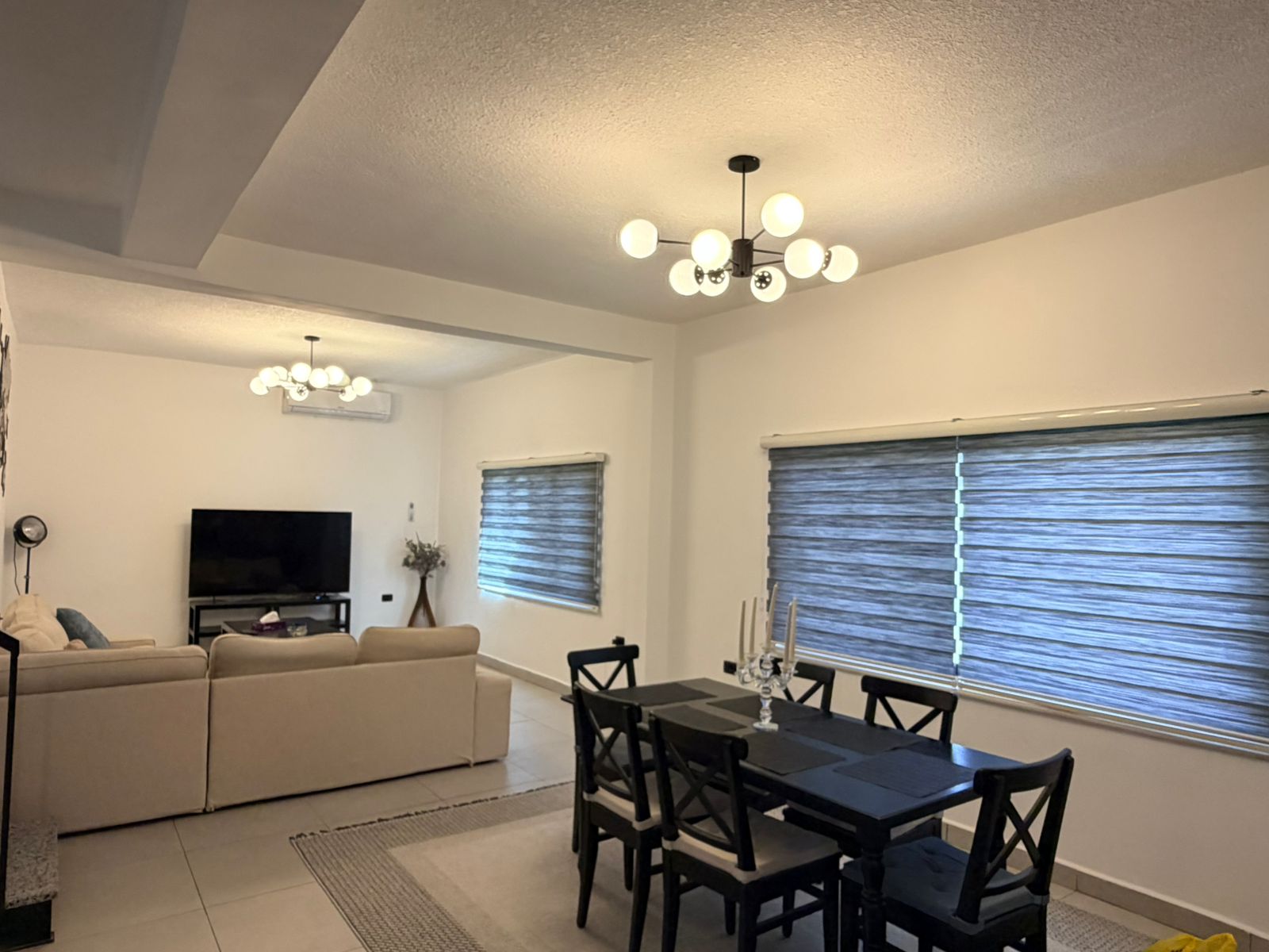 3d53958e-e0b6-4ebb-b72b-d2b7410a40d0 Open-concept living and dining room with a beige sectional, flat-screen TV on a stand, and large windows with gray striped blinds.