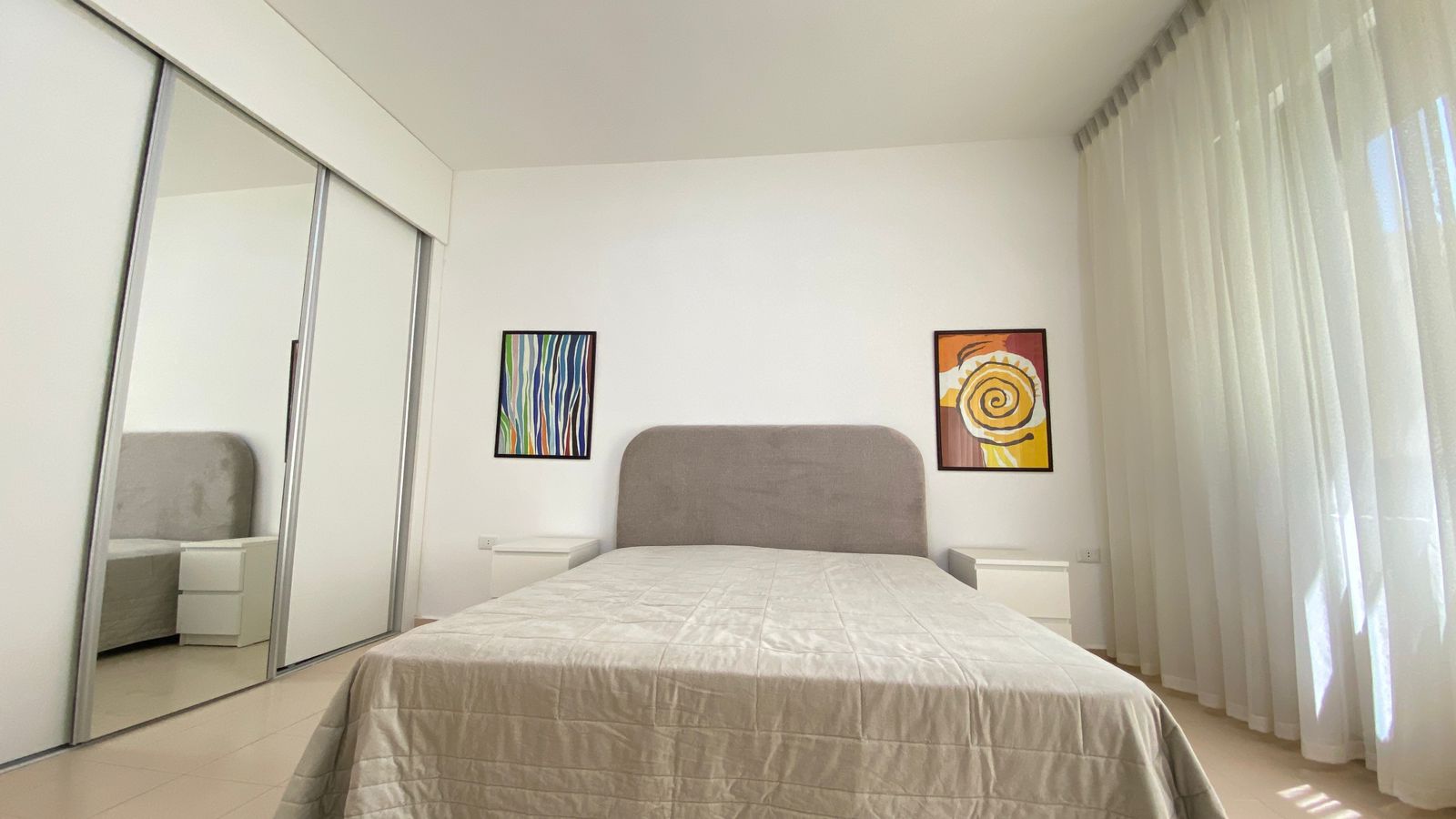 3e959e3d-e6b9-46cc-9e64-0915af52b1f2 Minimalist bedroom with a gray upholstered headboard, white bedding, and two white nightstands on either side of the bed.