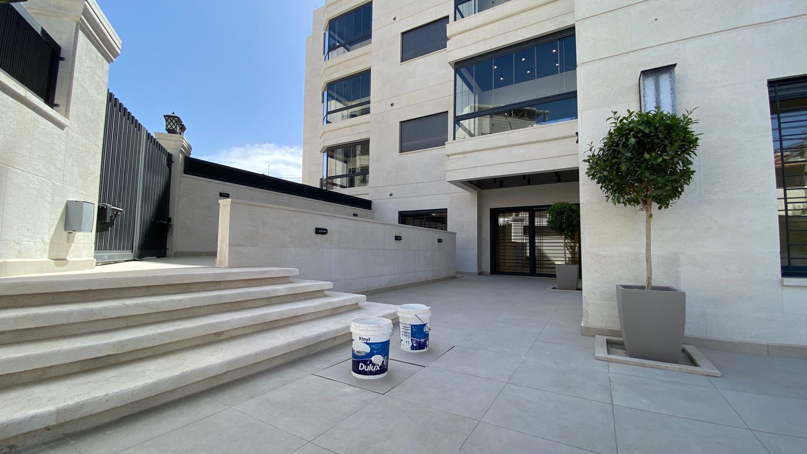 3eee1734-c77c-4a53-92c2-acd4f7e39e92 Modern white stone building entrance with a wide staircase, gated entry, and two paint buckets on the tiled plaza.