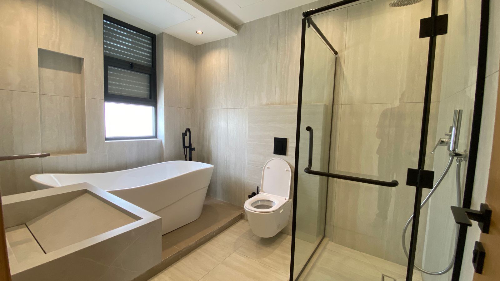 3f019687-7afb-4744-9390-f7ee0ef9ed6d Modern bathroom with a white freestanding tub on a raised platform, a wall-mounted black faucet, and a large window above. A wall-hung toilet sits next to a glass-enclosed shower with black metal framing and fixtures.