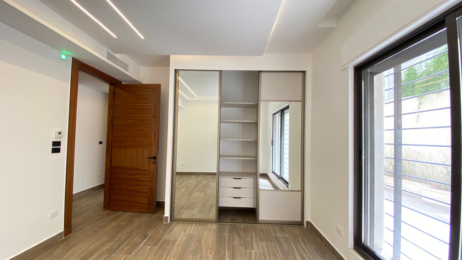 3f1371e9-02b6-41bd-bf05-23924f50cfba Modern bedroom with a built‑in wardrobe: sliding mirrored doors, open shelves and drawers, and a wooden entry door on the left; large window on the right.
