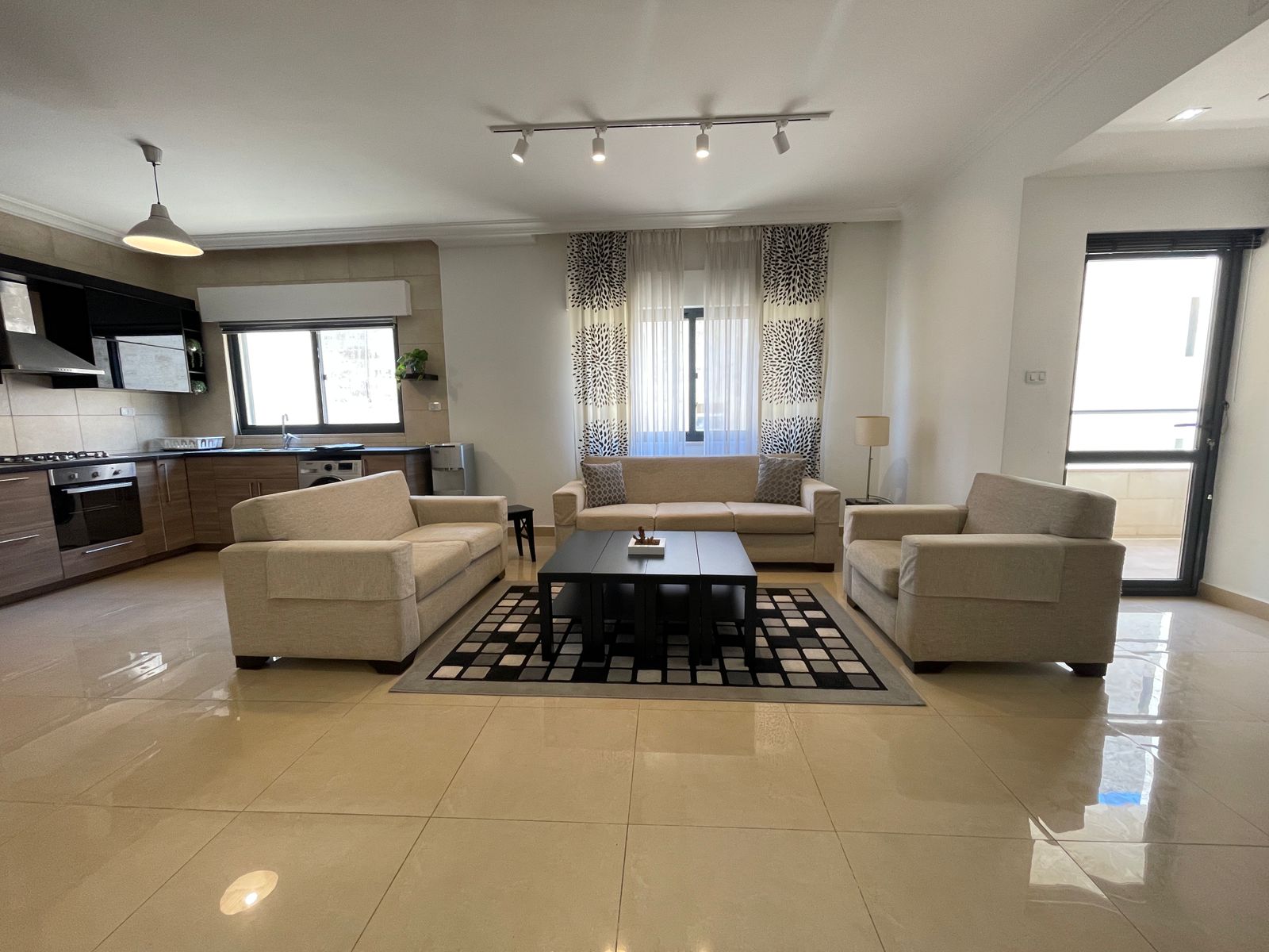3f93e873-bd60-41fb-84f3-e24705d0283e Open-plan living room with beige sofa set around a dark coffee table on a checker-pattern rug, adjacent to a modern kitchen.