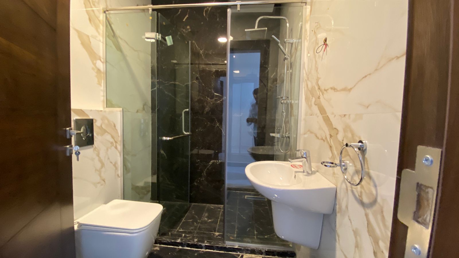 3f93e873-bd60-41fb-84f3-e24705d0283e Modern bathroom with a glass-enclosed shower, white toilet, and wall-mounted sink set against marble walls.