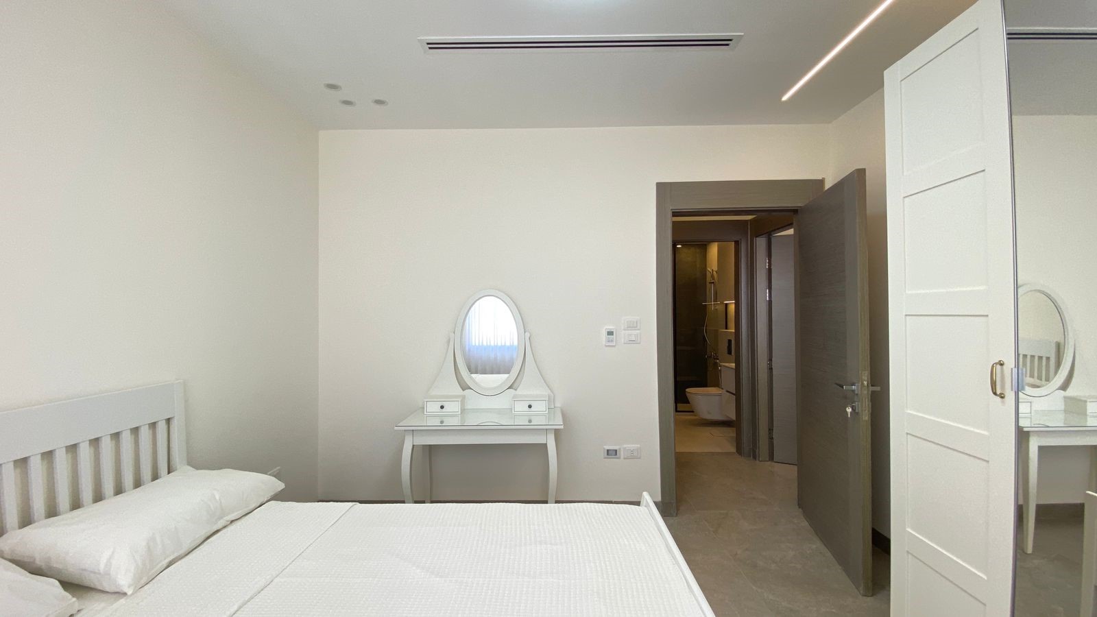 4083b486-b379-4325-8791-6fad1a4d57bd Bright white bedroom with a slatted white bed, a vanity dresser with oval mirror, and an open doorway to a bathroom.