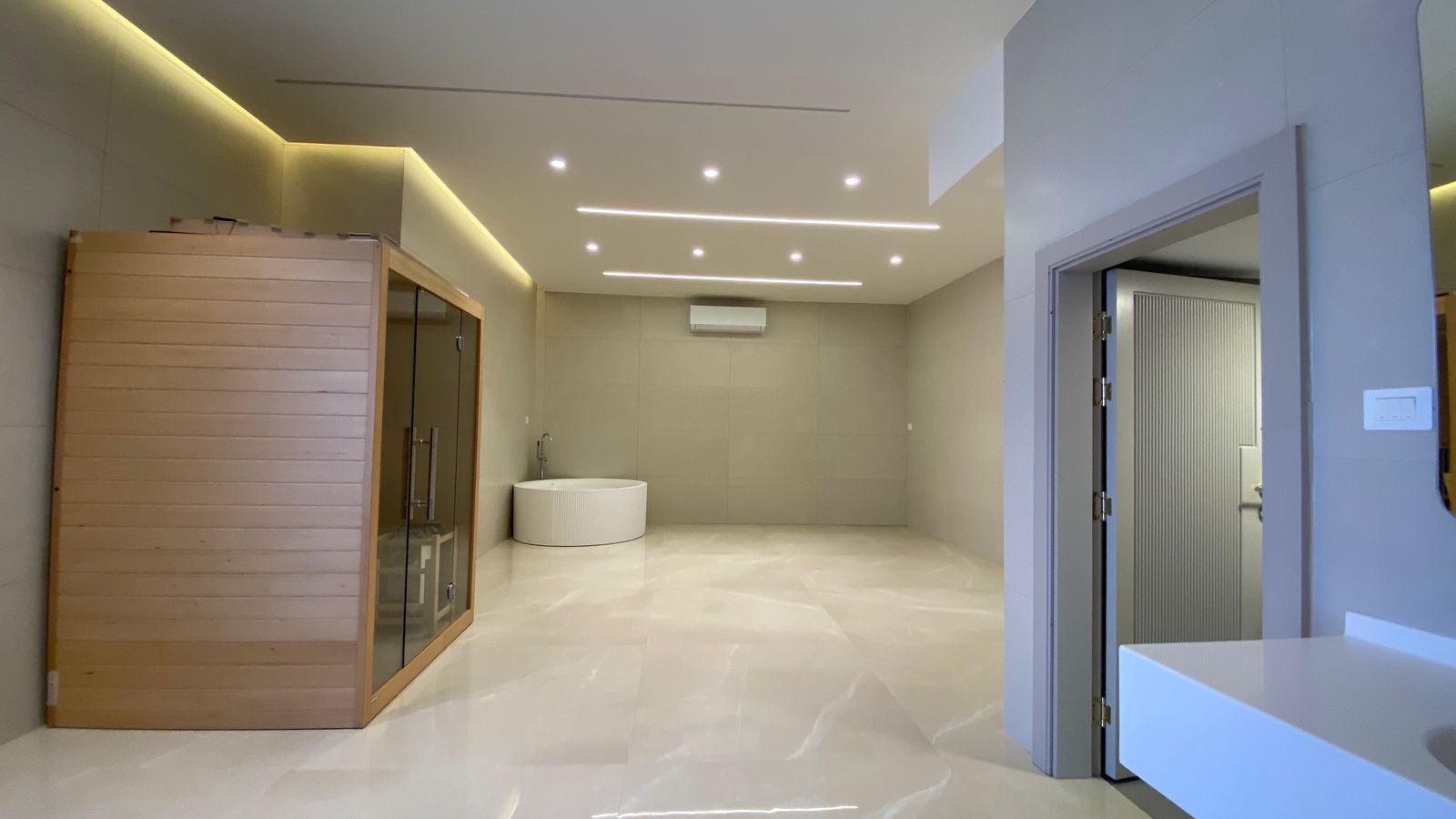 4161ae4a-5886-4e75-a3fd-3caa927aa01e Spa room with a wooden sauna on the left, a round white tub in the center, and a doorway on the right; glossy tiled floor.