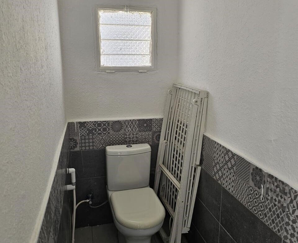 4178ea2c-4657-4dfd-999a-4ba75f48933b Small bathroom with a white toilet, patterned tile wainscoting, and a frosted square window above. A folded white radiator panel leans against the wall beside the toilet.