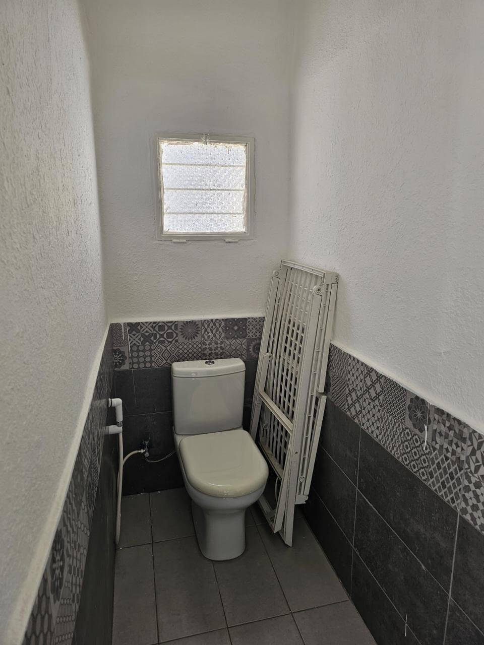 4178ea2c-4657-4dfd-999a-4ba75f48933b Small bathroom with a white toilet, patterned tile wainscoting, and a frosted square window above. A folded white radiator panel leans against the wall beside the toilet.