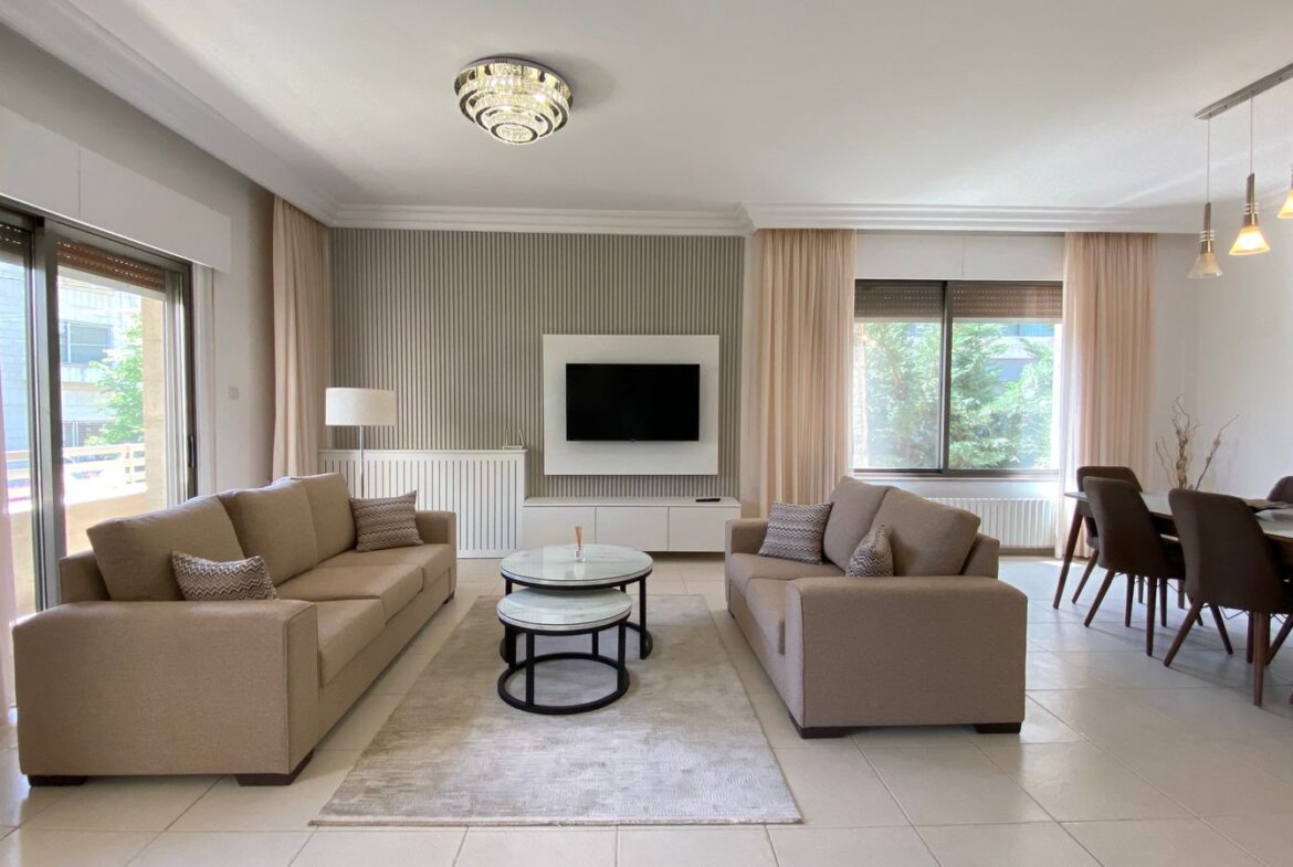 Bright open living room with two beige sofas facing a wall-mounted TV and marble-topped coffee tables on a light rug