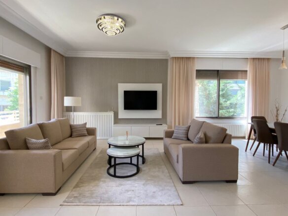 Bright open living room with two beige sofas facing a wall-mounted TV and marble-topped coffee tables on a light rug