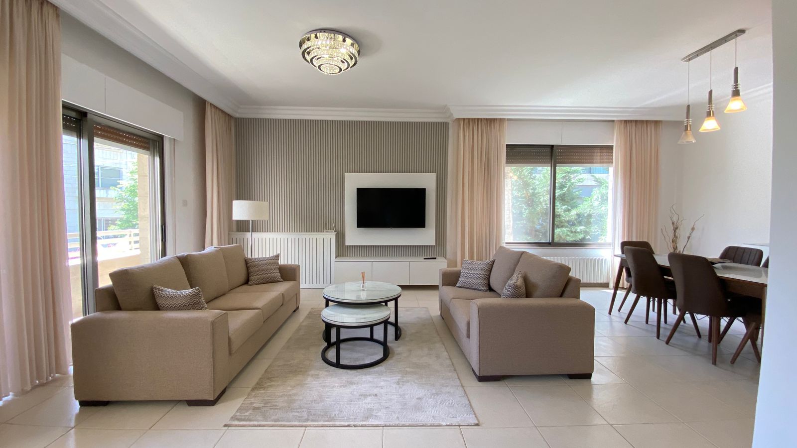 41dc5d66-3e50-4bf6-9a5f-784d2334e75c Bright open living room with two beige sofas facing a wall-mounted TV and marble-topped coffee tables on a light rug