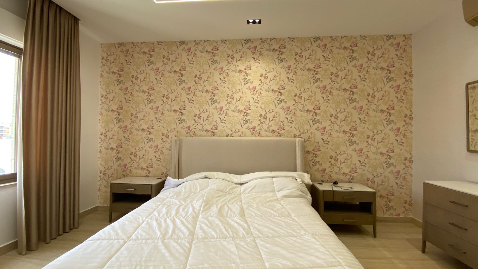 436b40e1-227f-4c57-b844-1aa5ba56fe63 Bedroom with a floral wallpaper feature wall, beige upholstered headboard, white quilted bed, and two matching nightstands.