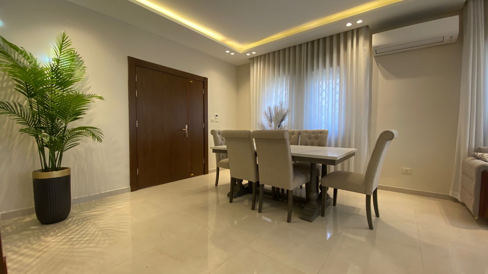 4486c285-ea1d-41c9-9b96-b5cd76a5f78c Modern dining room with a white table and beige upholstered chairs, a large potted palm, and sheer curtains.