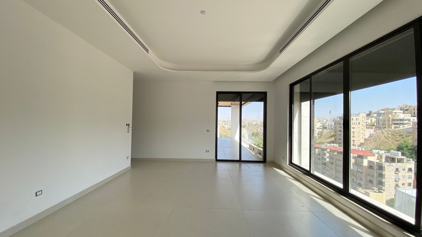 44d4b26e-536b-4591-a97d-26c8df33e9c1 Empty modern living room with large floor-to-ceiling windows and a balcony door, overlooking a cityscape.