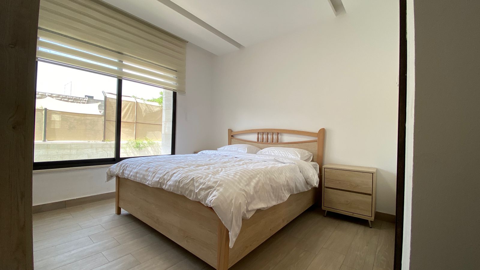 44dc9fb8-006d-483b-81c4-36909ffd4319 A wooden bed with white bedding and two pillows in a bright minimalist bedroom, beside a matching wooden nightstand near a large window with blinds.