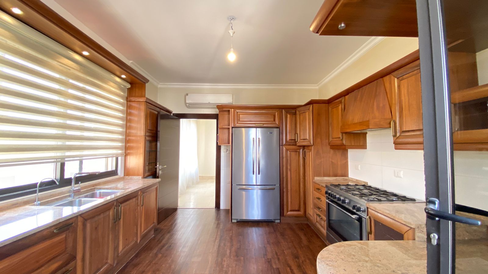 44e15f34-fb7a-481f-9ff7-c5930e891a6f Bright kitchen with dark wood cabinets, stainless fridge, and double sink by a wide window with blinds, and creamy counters visible at left.