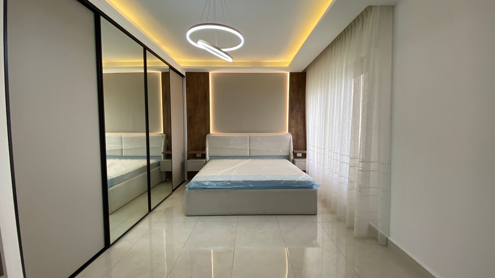 4517f2b4-93b0-4044-b7df-36de53611d70 Modern hotel bedroom with a low platform bed, backlit headboard panels, and a mirrored wardrobe on the left.