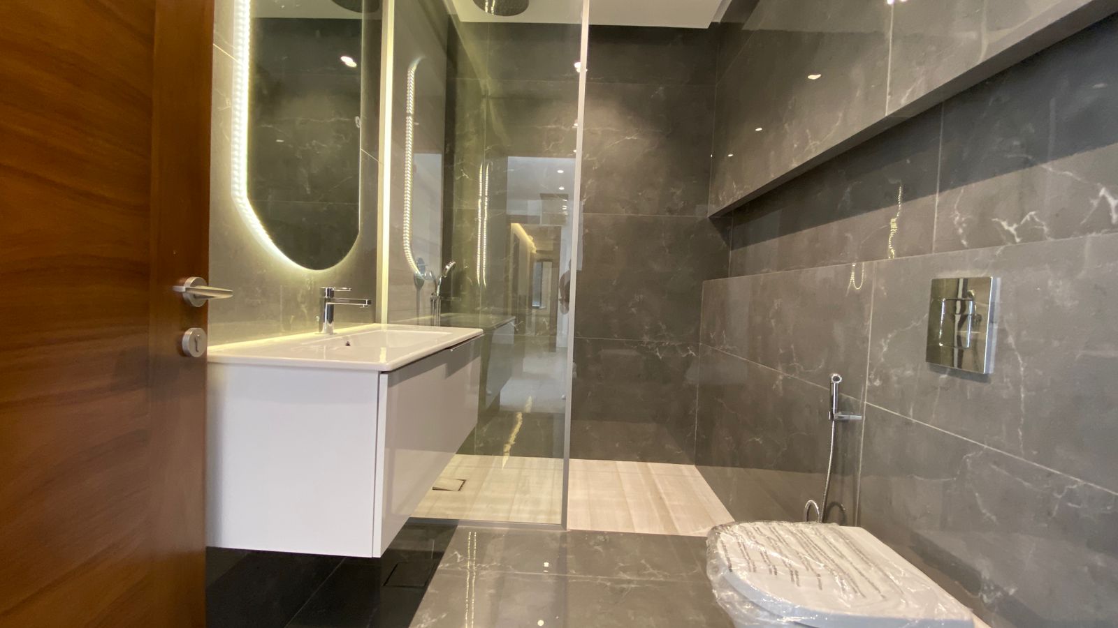 456d53d2-8a83-43eb-b775-959d4a3d812e Modern gray marble bathroom with a floating white vanity, backlit mirror, glass shower, and wall-mounted toilet.